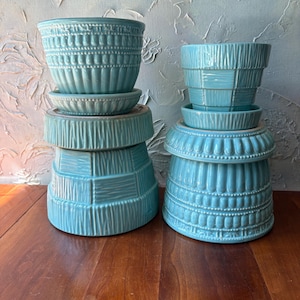 May include: Two stacks of turquoise ceramic plant pots with matching saucers. The left stack features a pot with vertical ribbed and beaded textures. The right stack includes a pot with a checkered pattern. Both sets have a glossy finish.