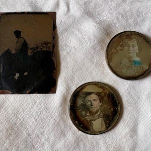 May include: A rectangular tintype photograph showing a silhouette of a person in a cap, alongside two circular tintype portraits. One circular portrait depicts a person with a blue accent on their clothing, and the other shows a person with a beard.