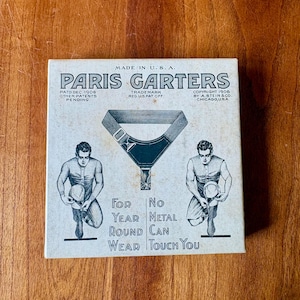 May include: A vintage off-white cardboard box for Paris Garters featuring black printed text and illustrations. The box displays two figures adjusting garters, the text "For Year Round Wear" and "No Metal Can Touch You," and patent details.