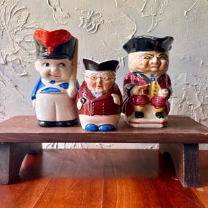 May include: Three ceramic character jugs featuring historical figures. The left jug wears a red and black hat with a blue coat. The center jug is a smaller figure in a dark coat and glasses. The right jug depicts a person in a black hat and red coat.