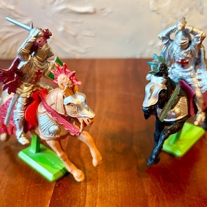 May include: Two toy knights on horseback. One knight is silver with red accents, holding a sword and shield. The other knight is silver with a red cross on the chest. Both are mounted on horses with green bases.