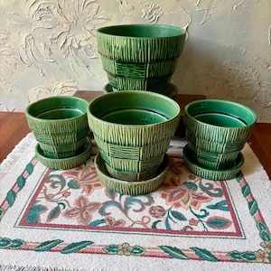 May include: A set of three green ceramic plant pots with a textured basket-weave pattern. Each pot features a matching circular saucer base. The pots are arranged with one larger pot in the back and two smaller pots in the front on a surface.