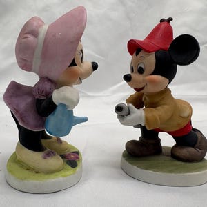 May include: Two ceramic figurines of cartoon characters. One figure is a mouse wearing a pink bonnet and dress, holding a blue watering can. The other figure is a mouse wearing a red hat and tan jacket, holding a camera. Both figures are on a round base.