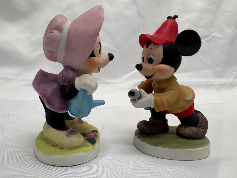 May include: Two ceramic figurines of cartoon characters. One figure is a mouse wearing a pink bonnet and dress, holding a blue watering can. The other figure is a mouse wearing a red hat and tan jacket, holding a camera. Both figures are on a round base.