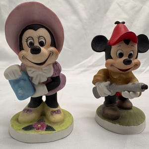 May include: Two ceramic figurines of Disney characters. Minnie Mouse wears a pink hat and purple dress, holding a blue item. Mickey Mouse wears a red hat and brown shirt, holding a gray toy gun. Both stand on a round base.