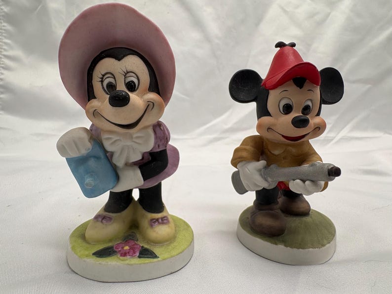 May include: Two ceramic figurines of Disney characters. Minnie Mouse wears a pink hat and purple dress, holding a blue item. Mickey Mouse wears a red hat and brown shirt, holding a gray toy gun. Both stand on a round base.