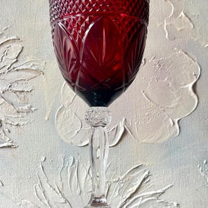 May include: A ruby red wine glass with a clear stem and base. The bowl features a diamond and leaf pattern. The glass sits on a wooden surface against a textured white and blue background.
