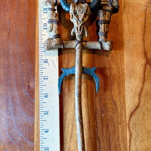 May include: A detailed, decorative staff with a skull and horns design. The staff is approximately 15 inches long, featuring a cross-shaped top with a skull, blue horns, and a clear orb. The staff has a wooden appearance with blue accents.