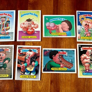 May include: A collection of eight Garbage Pail Kids trading cards. The cards feature various grotesque characters with humorous names like "Roy L. Flush" and "Frank Footer." The cards are on a wooden surface.
