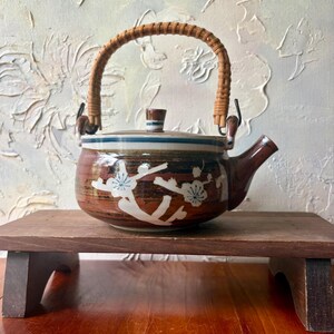 May include: A ceramic teapot with a brown and white glaze featuring a painted white floral branch design. The teapot has a rounded body, a short spout, a matching lid with a small knob, and a curved, woven rattan handle attached to metal loops.