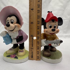 May include: Two ceramic figurines of cartoon characters. One is a female character wearing a pink hat and dress, holding a blue object. The other is a male character wearing a red hat and holding a gray object. The figurines are approximately 4 inches tall.