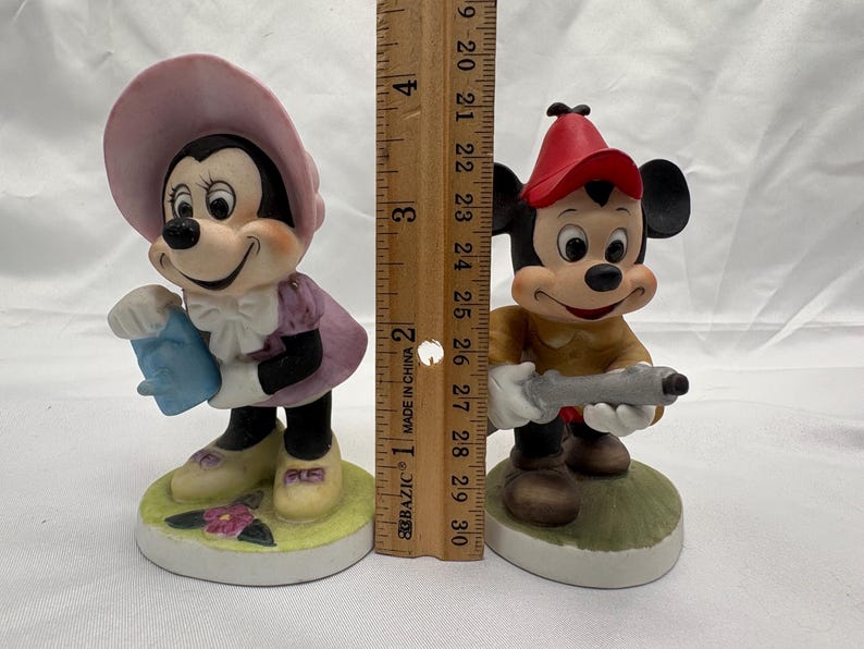 May include: Two ceramic figurines of cartoon characters. One is a female character wearing a pink hat and dress, holding a blue object. The other is a male character wearing a red hat and holding a gray object. The figurines are approximately 4 inches tall.