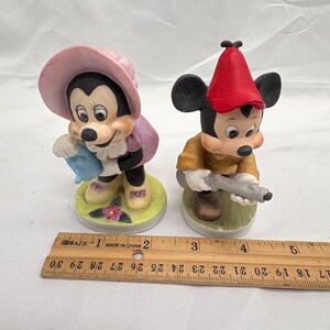May include: Two ceramic figurines of Mickey and Minnie Mouse. Minnie wears a pink hat and purple dress, holding a blue item. Mickey wears a red hat and yellow shirt, holding a gray object. Both stand on circular bases.