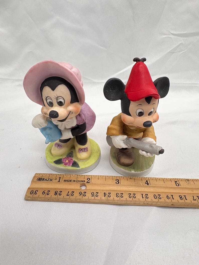May include: Two ceramic figurines of Mickey and Minnie Mouse. Minnie wears a pink hat and purple dress, holding a blue item. Mickey wears a red hat and yellow shirt, holding a gray object. Both stand on circular bases.