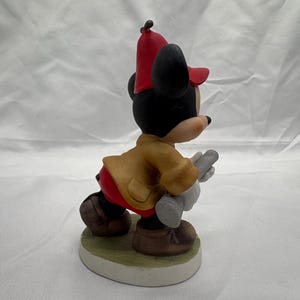 May include: A ceramic figurine of Mickey Mouse in a fireman's outfit. Mickey is wearing a red hat, tan jacket, red pants, and brown shoes. He is holding a gray object and is standing on a green and white base.
