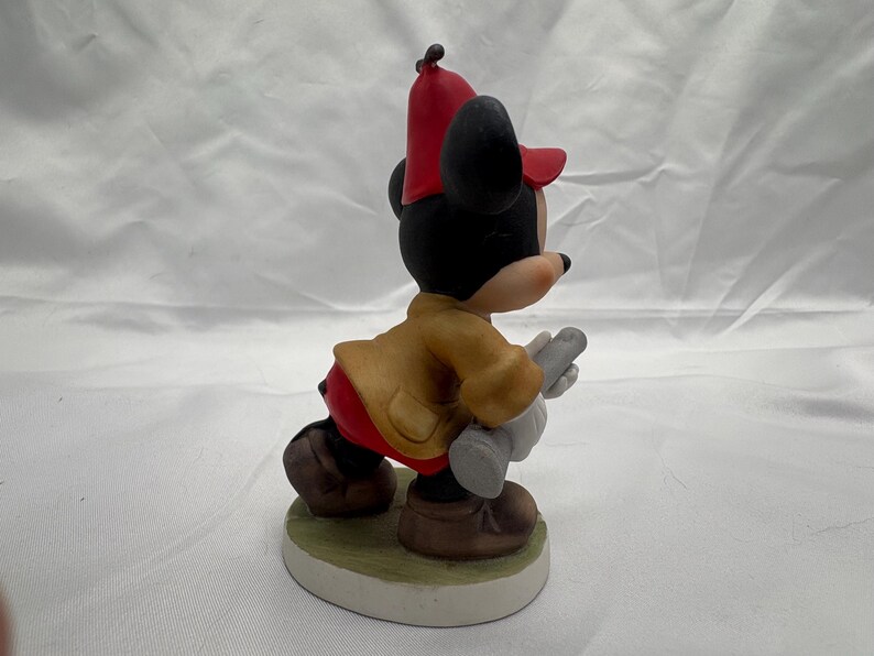 May include: A ceramic figurine of Mickey Mouse in a fireman's outfit. Mickey is wearing a red hat, tan jacket, red pants, and brown shoes. He is holding a gray object and is standing on a green and white base.
