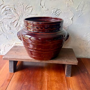 May include: A dark brown glazed ceramic crock featuring a wide mouth, two side handles, and a decorative embossed pattern of horizontal wavy lines and small flower motifs encircling the exterior of the rounded, sturdy vessel body.