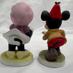 May include: Two ceramic figurines of cartoon characters. One figure is wearing a pink hat and purple dress, the other is Mickey Mouse in a red hat, tan jacket, and red pants. Both figures are on a round base.