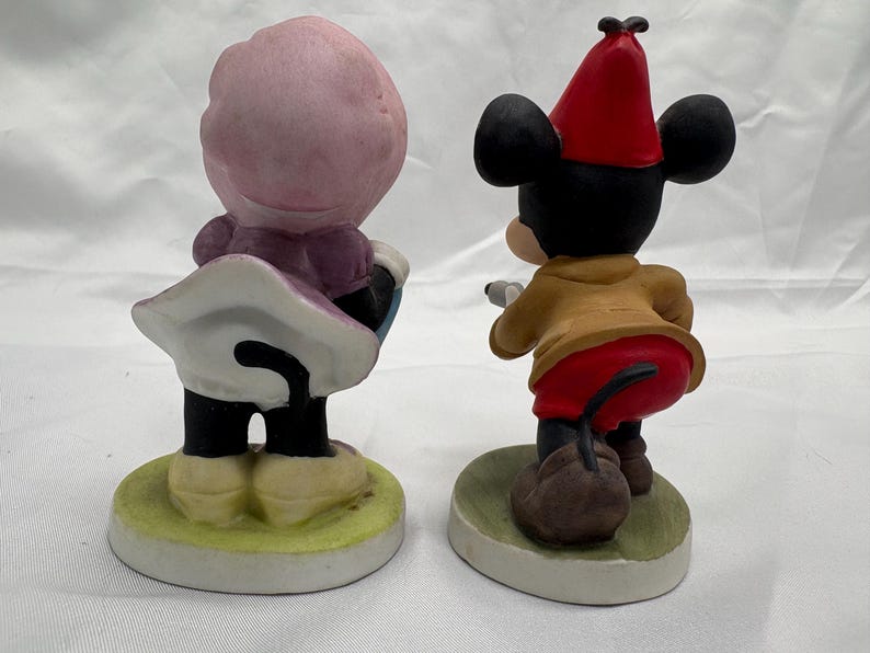 May include: Two ceramic figurines of cartoon characters. One figure is wearing a pink hat and purple dress, the other is Mickey Mouse in a red hat, tan jacket, and red pants. Both figures are on a round base.