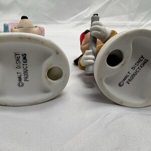 May include: Two ceramic figurines of Disney characters, viewed from the bottom. One figurine features a mouse with pink ears and a blue hat. The other figurine is of a mouse holding a gray object. Both figurines have a white base with the text "WALT DISNEY PRODUCTIONS".