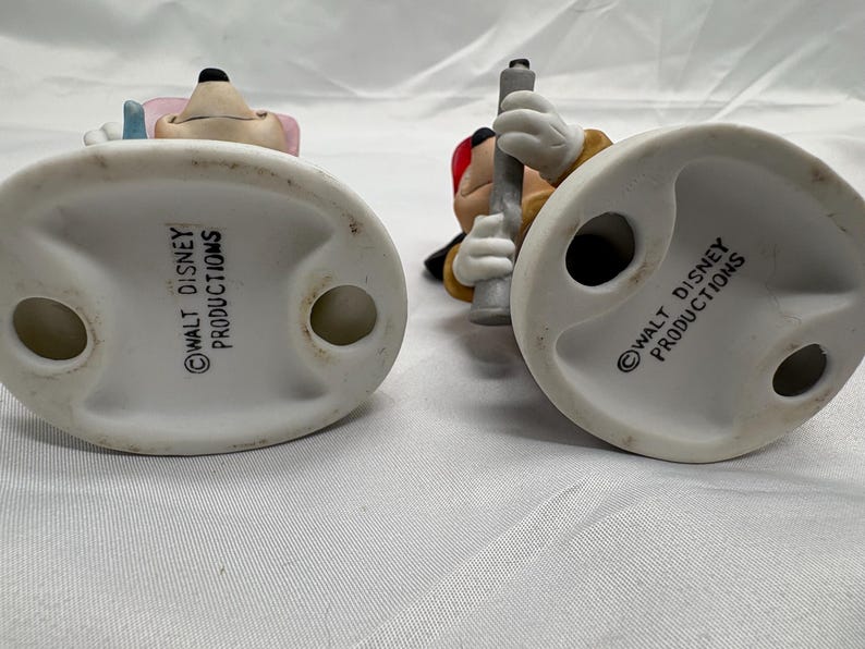May include: Two ceramic figurines of Disney characters, viewed from the bottom. One figurine features a mouse with pink ears and a blue hat. The other figurine is of a mouse holding a gray object. Both figurines have a white base with the text "WALT DISNEY PRODUCTIONS".