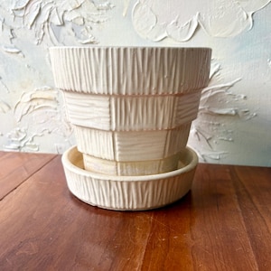 May include: A cream-colored ceramic plant pot with a matching saucer, featuring a tiered, stepped silhouette and a vertical, textured, ribbed pattern across the exterior surface of both the pot and the base, creating a consistent, repeating design.