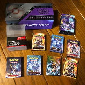 May include: A collection of Pokemon trading cards, including booster packs and a trainer's toolkit. The booster packs feature various Pokemon from the Sun & Moon series, including Burning Shadows and Guardians Rising. The trainer's toolkit includes a toploader combo box.