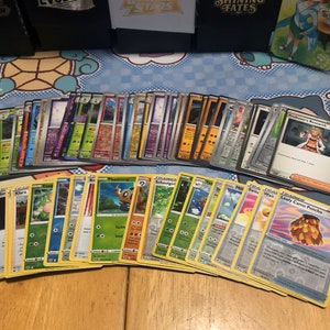 May include: A collection of Pokemon trading cards featuring various characters and abilities. The cards are arranged in rows on a wooden table. Some of the cards include "Professor's Research", "Klara", "Phantump", "Schoolgirl", "Milt", "Switc", "Wait", and "Leafy Camo Poncho".