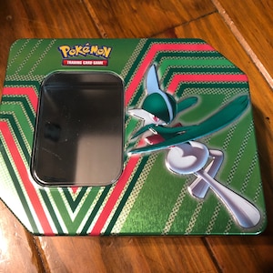 Puede incluir: Lata de metal con rayas verdes y rojas con un logotipo de Pokemon Trading Card Game y una imagen de un personaje Pokemon verde y blanco.