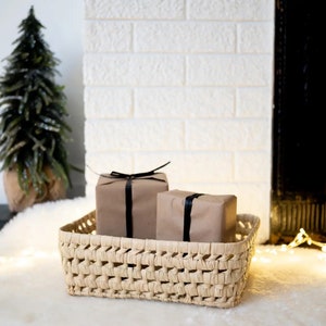 May include: A woven wicker basket with two wrapped gifts inside. The gifts are wrapped in brown paper and tied with black ribbon. The basket is sitting on a white fluffy surface.
