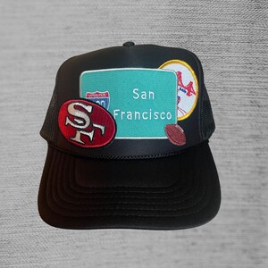 May include: Black trucker hat with a green road sign that says "San Francisco" and a red and white "SF" logo. The hat also has a red, white, and blue baseball logo and a brown football.