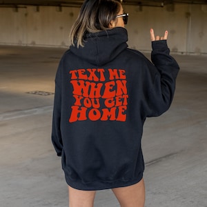 K&ouml;nnte beinhalten: Schwarzer Kapuzenpullover mit dem Text "TEXT ME WHEN YOU GET HOME" in roter Retro-Schrift auf der R&uuml;ckseite.