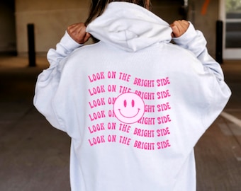 May include: A light blue hoodie with the phrase "LOOK ON THE BRIGHT SIDE" printed in pink. The design includes a smiling face graphic. The hoodie is a casual garment, suitable for everyday wear.