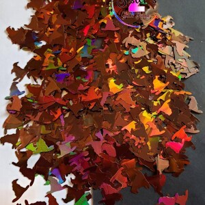 May include: A pile of iridescent brown witch hat shaped glitter. The glitter is scattered on a white surface and a black surface.