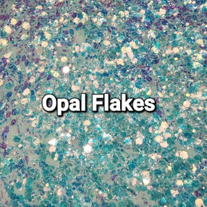 May include: A close-up of iridescent blue and white glitter flakes. The glitter is scattered randomly across the surface, creating a shimmering effect. The text "Opal Flakes" is printed in white letters on the glitter.