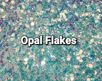 May include: A close-up of iridescent blue and white glitter flakes. The glitter is scattered randomly across the surface, creating a shimmering effect. The text "Opal Flakes" is printed in white letters on the glitter.
