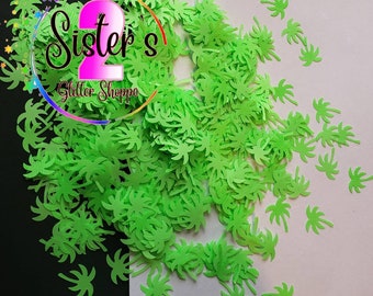 May include: A large pile of green palm tree shaped glitter. The glitter is on a white surface with a black background. The text "Sister's Glitter Shoppe" is visible in the image.