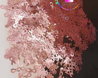 May include: A large pile of rose gold glitter in the shape of awareness ribbons. The glitter is scattered on a white surface and a black surface. The text "Sister's Glitter Shoppe" is visible in the image.