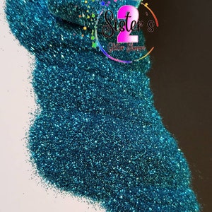 May include: Teal glitter with a fine, shimmery texture. The glitter is loose and scattered on a white surface. The text "Sister's Glitter Shoppe" is visible in the upper right corner of the image.