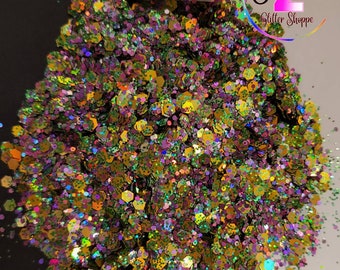 May include: A close-up of iridescent glitter in shades of green, gold, and purple. The glitter is scattered on a white surface. The text "Sister's Glitter Shoppe" is visible in the upper right corner of the image.