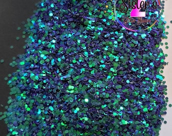 May include: A close-up of a pile of iridescent glitter in shades of blue and green. The glitter is hexagonal in shape and appears to be spilling from a white container. The logo "Sister's Glitter Shoppe" is visible.