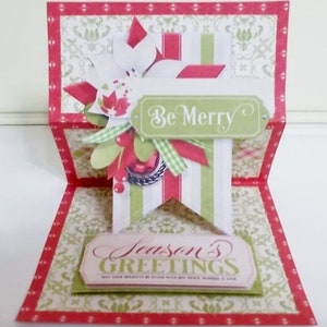 May include: A red, white, and green Christmas card with a pop-up design. The card features a banner with the words "Be Merry" and a smaller banner with the words "Season's Greetings".