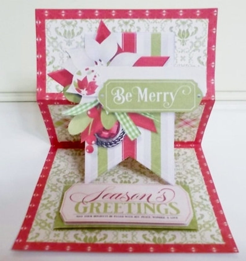 May include: A red, white, and green Christmas card with a pop-up design. The card features a banner with the words "Be Merry" and a smaller banner with the words "Season's Greetings".
