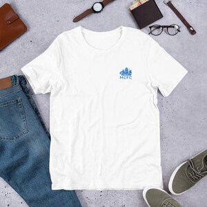 以下が含まれることがあります： 左胸に青い船のロゴと「MCFC」の文字が入った白いTシャツです。