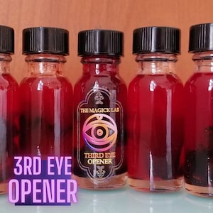 Puede incluir: Cinco pequeños frascos de vidrio con tapas negras llenos de un líquido rojo. La etiqueta en el frasco del centro dice "The Magick Lab" y "Third Eye Opener".