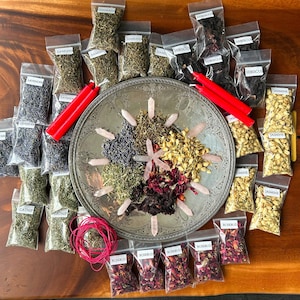 Puede incluir: Una bandeja plateada con varias hierbas y flores secas en pequeñas bolsas de plástico. Las hierbas y las flores están dispuestas en círculo alrededor de la bandeja. También hay dos velas rojas y un carrete de cuerda roja en la bandeja.