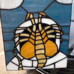 May include: Stained glass panel featuring a stylized butterfly design. The artwork incorporates white, yellow, orange, and black elements, set against a blue background with two white moon shapes. The panel is framed with dark lines.