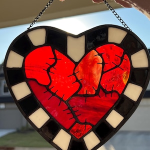 May include: A heart-shaped stained glass panel with a red centre, black stitching, and a black, white, and black border. It hangs from a black chain. The design evokes a broken heart.