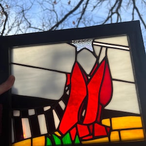 May include: Stained glass art piece with a black frame. The artwork features a vibrant red design against a white and gray background, with accents of yellow, green, and black. The piece is set against a backdrop of a blue sky and tree branches.