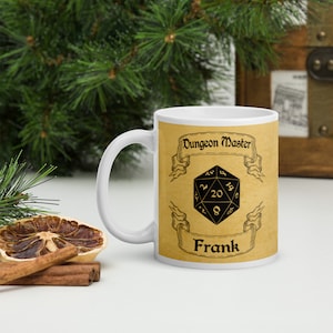 Könnte beinhalten: Weiße Keramiktasse mit goldfarbenem Hintergrund und schwarzem Text. Der Text lautet "Dungeon Master" mit einem Banner und einem schwarzen 20-seitigen Würfel. Der Name "Frank" steht unter dem Würfel.